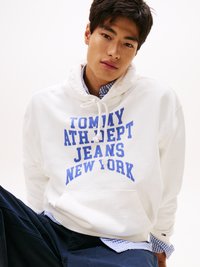 Balta kapuce ar zilu tekstu "TOMMY ATH.DEPT JEANS NEW YORK", ar auklu kapuci un priekšējo kabatu, uzvilkta virs zila rūtotā krekla.