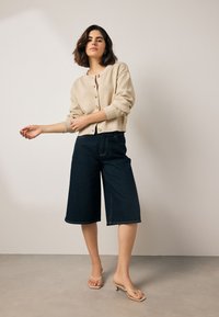 Cardigan beige en tricot avec encolure ronde et boutons dorés, associé à un short en jean bleu marine à jambe large et des tongs couleur tan.