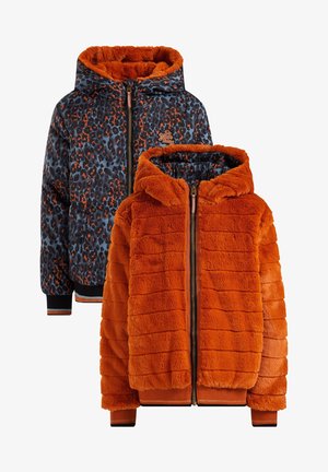 Persona con una chaqueta de felpa naranja brillante con forro de estampado de leopardo, vaqueros azul claro, camiseta negra y botas azules con cordones.