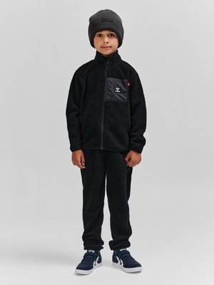 Kind draagt een zwarte fleecejas en broekset met een grijze muts en marineblauwe sneakers, staand tegen een effen grijze achtergrond.