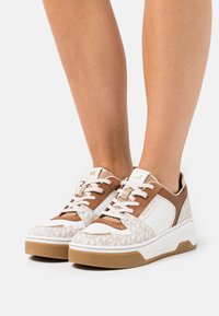 Sneaker bianchi e beige con parte superiore testurizzata, caratterizzate da un design con logo, collo imbottito e chiusura con lacci. Suola in gomma marrone chiaro.