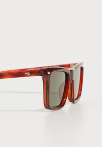 Gafas de sol de plástico tintado en rojo con montura cuadrada, lentes gris oscuro y pequeños detalles en dorado en las patillas.