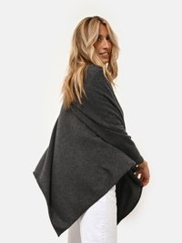 Poncho envolvente en gris carbón de tejido suave, con un ajuste holgado y bordes asimétricos que le dan un aspecto texturizado. Combinado con pantalones blancos.