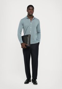 Camisa de manga longa azul clara, abotoada com colarinho, combinada com calças escuras e sapatos pretos. A segurar uma carteira preta texturizada.