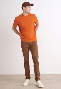 Oransje kortermet skjorte, brune slim-fit bukser og hvite sneakers. Tilbehør inkluderer en brun cap. Enkel design uten synlige mønstre.