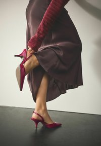 Escarpins à bride arrière en cuir verni rouge avec bout pointu et talon fin, portés avec une jupe en soie foncée légèrement brillante.