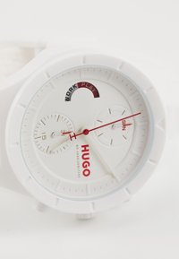 White watch with chronograph dials, red accents, and “HUGO” branding. Features a smooth silicone band and clear glass over a textured face.
