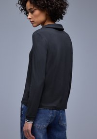 Haut noir à manches longues avec col, caractérisé par une texture lisse et des accents rayés aux poignets, associé à un jean en denim bleu.