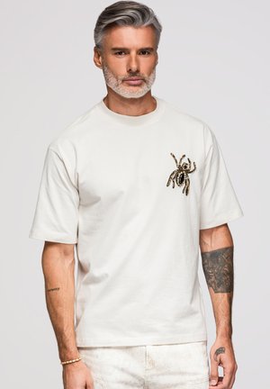 Mand med gråt hår og skæg bærer en hvid t-shirt med et stort edderkopdesign på brystet og har tatoveringer på begge underarme.