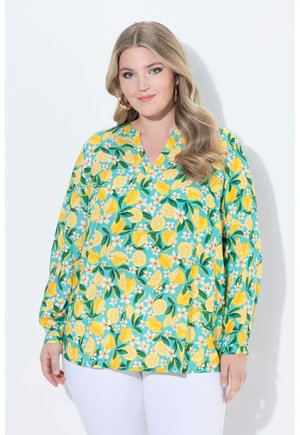 Frau mit langen, welligen Haaren, die goldene Creolen-Ohrringe und eine hellfarbige Bluse mit Zitronen- und Blumenmuster trägt, kombiniert mit weißen Hosen.