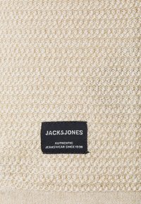 Tissu beige tricoté avec un motif texturé, avec une étiquette noire portant un texte blanc indiquant "JACK & JONES AUTHENTIC JEANSWEAR SINCE 1990."