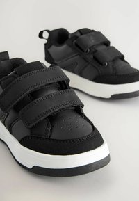 Baskets noires en cuir et en daim, dotées de trois bandes réglables en Velcro, d'une semelle intermédiaire blanche solide et d'une semelle extérieure texturée noire.