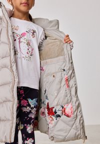 Párnázott szürke mellény virágos béléssel és arany kiegészítőkkel, puha belső résszel, cipzáros zárással és logóval a bélésen. Grafikás pólóval és mintás leggings-szel párosítva.