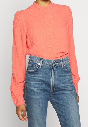 Person trägt eine korallenrosa strukturierte Bluse mit Knöpfen und langen Ärmeln, die in eine hoch taillierte blaue Jeans aus Denim gesteckt ist.