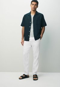 Short-sleeve dark green shirt with button front, white linen pants, and black sandals. The shirt has a chest pocket and relaxed fit.