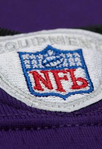 Besticktes NFL-Patch mit einem blauen Schild, das von Weiß umrandet ist, rotem "NFL"-Text auf einem purpurfarbenen Stoffhintergrund. Die Textur wirkt weich.