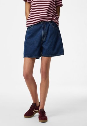 Personne portant un short en denim bleu, un t-shirt rayé bordeaux et blanc, et des baskets bordeaux avec des accents beige, debout devant un fond uni.