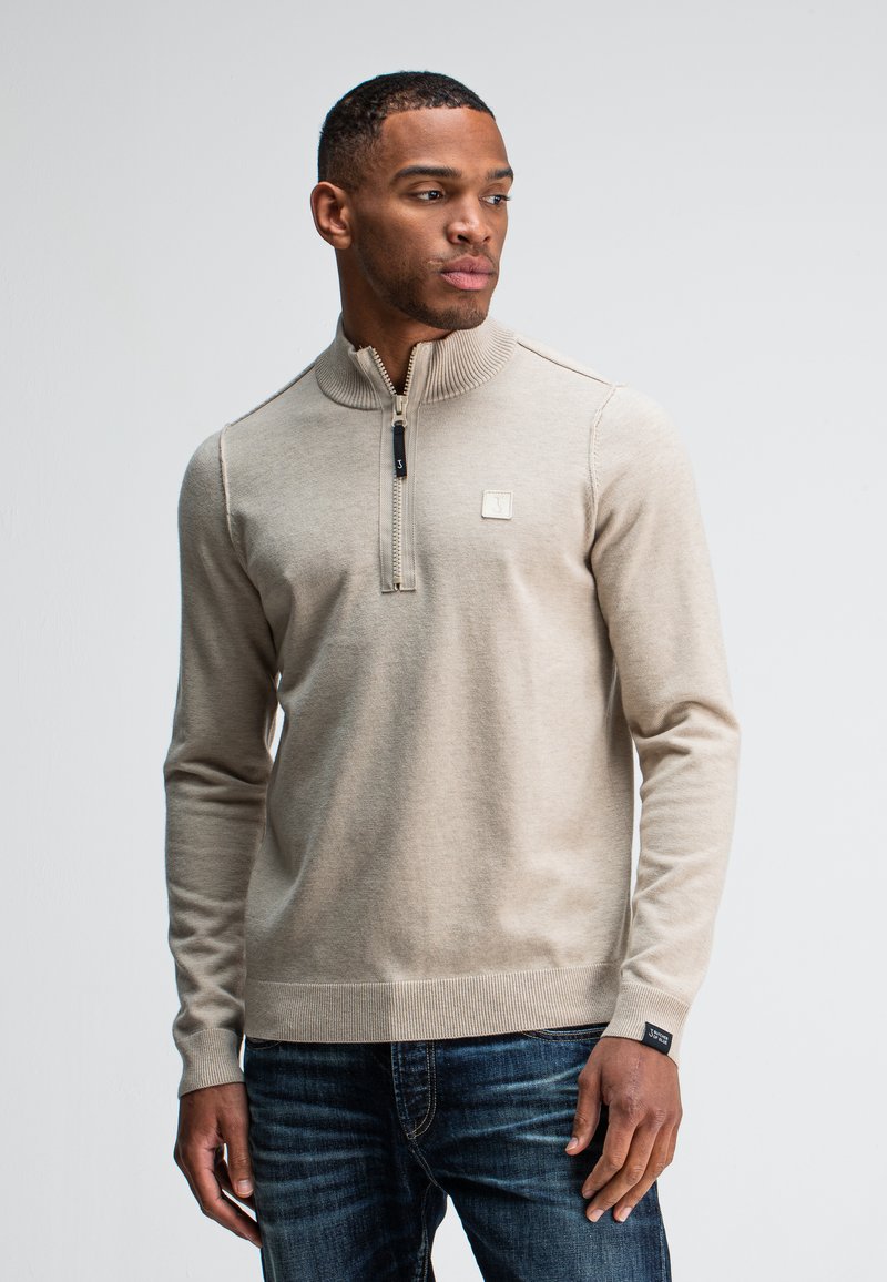 Beige ritsvest met een hoge ribbelkraag, textuurstof en een klein logo-opdruk. Draag het over blauwe jeans met zichtbare stiksels.