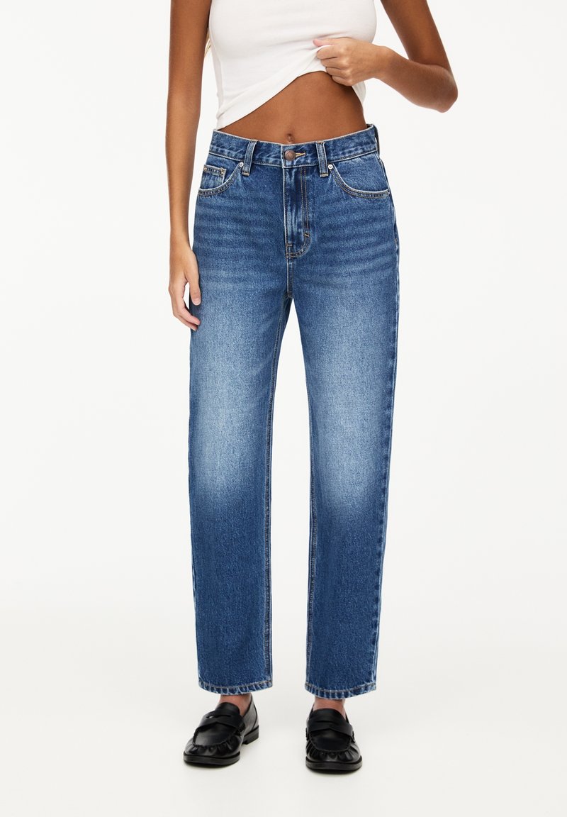 Femme portant un jean bleu taille haute à jambes droites, un top court blanc et des mocassins noirs à enfiler, debout devant un fond uni.