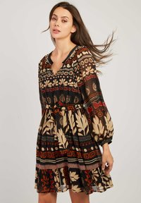 Femme avec de longs cheveux bruns portant une robe à motifs ceinturée dans des tons terreux et des manches longues en tulle, les cheveux soufflés sur le côté devant un fond uni.