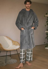 Homme debout pieds nus portant un peignoir gris texturé sur un pantalon de pyjama à carreaux dans un cadre intérieur chaleureux avec une chaise et des plantes.