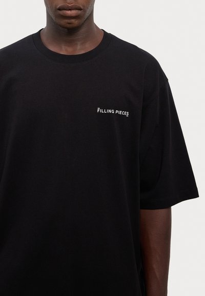 Svart bomullströja med rund halsringning och korta ärmar. Framsidan visar varumärket "Filling Pieces" i vit text.