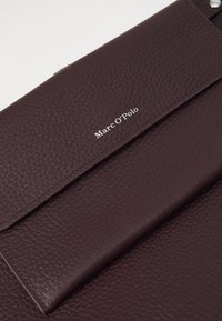 Pochette en cuir bordeaux avec une surface texturée, dotée d'un rabat avant et d'un logo "Marc O’Polo" en relief de couleur blanche. Accents en métal argenté.
