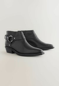 Botines de cuero negro hasta el tobillo con punta afilada, cierre de cremallera y una correa decorativa con detalles de hebilla de metal. Tacón bajo apilado.