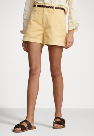 Person iført gule shorts med flettet brunt bælte, en lys stribet skjorte og mørkebrune åbne sandaler, der står mod en ensfarvet baggrund.