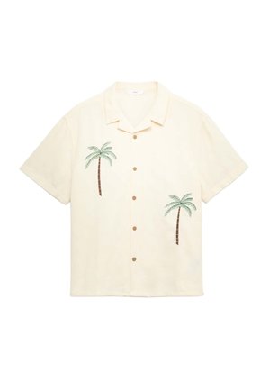 Ung person står med hænderne i lommerne iført en cremegul kortærmet skjorte med palmeprint, hvid t-shirt, grønne shorts og sneakers.