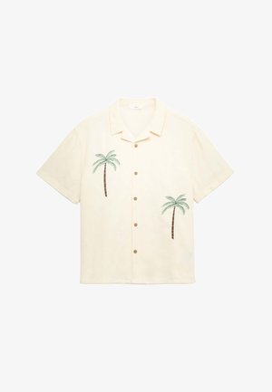 Junge Person steht mit den Händen in den Taschen, trägt ein cremefarbenes kurzärmeliges Hemd mit Palmenmuster, ein weißes T-Shirt, grüne Shorts und Sneakers.