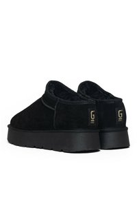 Zapatos de ante negro con un interior acolchado, forma redondeada y suela gruesa de goma negra. Presentan una etiqueta de marca en el talón.