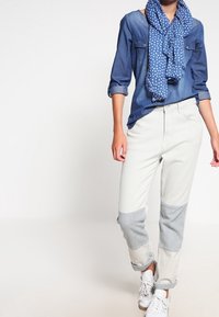 Camicia di jeans a maniche lunghe, sciarpa azzurra chiara con pois bianchi, pantaloni a blocchi di colore grigio chiaro e blu e sneakers bianche.