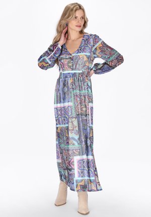 Lyshåret kvinde iført en langærmet, V-hals maxikjole med farverige, geometriske og blomsteragtige mønstre, kombineret med beige ankelstøvler.