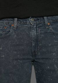 Gros plan d'un jean en denim noir délavé avec fermeture par boutons, passants de ceinture et poches avant, porté avec une chemise noire.