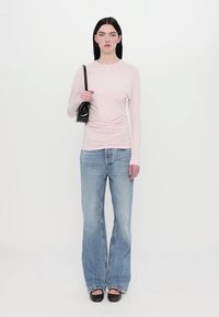Light pink long-sleeve top with gathered detail, paired with light blue flared jeans and black handbag. Model stands against a white background.
