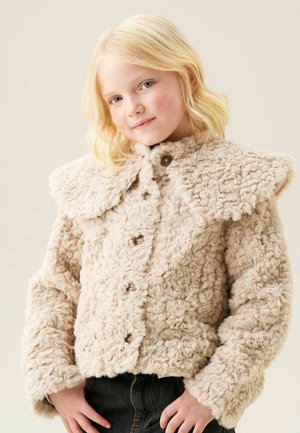 Lille barn med blondt hår iført en beige, fluffyjakke med knapper og mørke bukser, smilende let med hænderne hvilende nær hofterne.