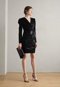 Robe noire à sequins avec un décolleté en V, de longues manches ballon et des détails froncés. Portée avec des escarpins noirs à lanières et une pochette noire slim.