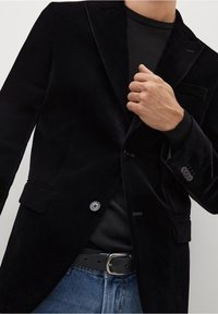Persona con giacca di velluto nero sopra una camicia nera e jeans blu con cintura nera, che aggiusta il risvolto della giacca con la mano destra.