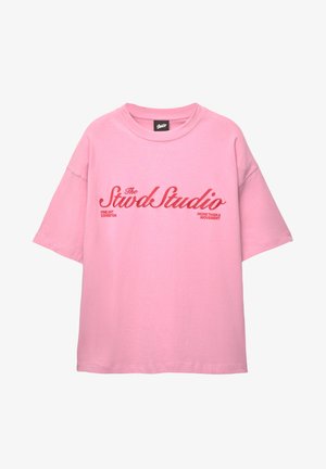 Vaaleanpunainen lyhythihainen puuvillapaita, jossa on punaisella tekstillä kirjoitettu "The Stwd Studio" ja "Fine Art Exhibition", löysällä, rentouttavalla istuvuudella.