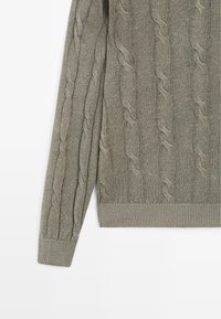 Manche de pull tricotée marron clair avec motif torsadé et poignets et ourlet côtelés sur fond blanc.