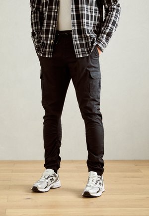 Person wearing black cargo jogger pants, black and white plaid shirt, and white New Balance sneakers standing on wooden floor.