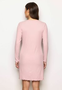 Femeie îmbrăcată într-o rochie lungă, de culoare roz deschis, cu mâneci lungi și lungime până la genunchi, cu o margine în valuri, stând împotriva unui zid alb simplu, întoarsă cu spatele.