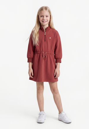 Jeune fille aux longs cheveux blonds portant une robe rouge à manches longues avec une fermeture éclair devant et des baskets blanches, debout et souriante.