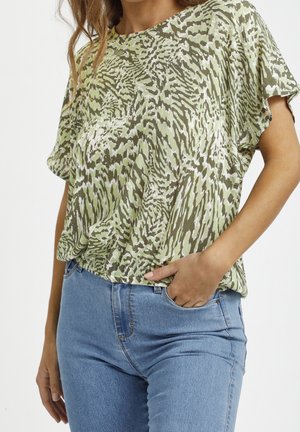 Kobieta w zielono-białej bluzce z krótkim rękawem o abstrakcyjnym wzorze, wpuszczonej w jasnoniebieskie dżinsy z wysokim stanem, z ręką w kieszeni.