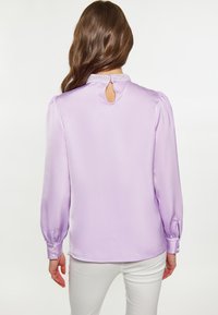 Lavendelfarbene Satinbluse mit puffärmeligen langen Ärmeln, einem Schlüsselloch-Rücken und verzierten Kragen. Sie hat Knopfdetails an den Bündchen.
