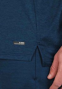 Marineblauwe sportshirt met een gestructureerde stof, zijsplit en een JAKO-merktag nabij de zoom. Het heeft een ontspannen pasvorm.