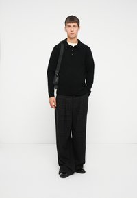 Schwarzer gestrickter Pullover mit Kragen, kombiniert mit schwarzer und grauer karierten weit geschnittenen Hosen, schwarzer Schultertasche und glänzenden schwarzen Schuhen.