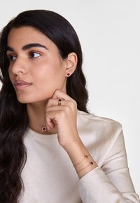 Bijuterii cu ton de aur, având pietre prețioase circulare roșii. Include cercei tip studs, un inel și brățări stratificate cu modele asortate, toate purtate pe un top deschis la culoare.