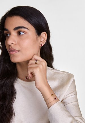 Bijuterii cu ton de aur, având pietre prețioase circulare roșii. Include cercei tip studs, un inel și brățări stratificate cu modele asortate, toate purtate pe un top deschis la culoare.